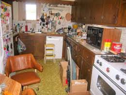 Photo of a cluttered, outdated kitchen, all counterspace filled, all cabinets old and falling apart.
