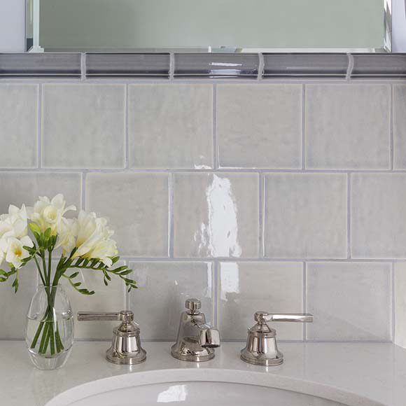 Photo of bathroom detail with white tiling behind a white sink, silver handles, flowers in a vase, and a mirror above a single line of grey tile.