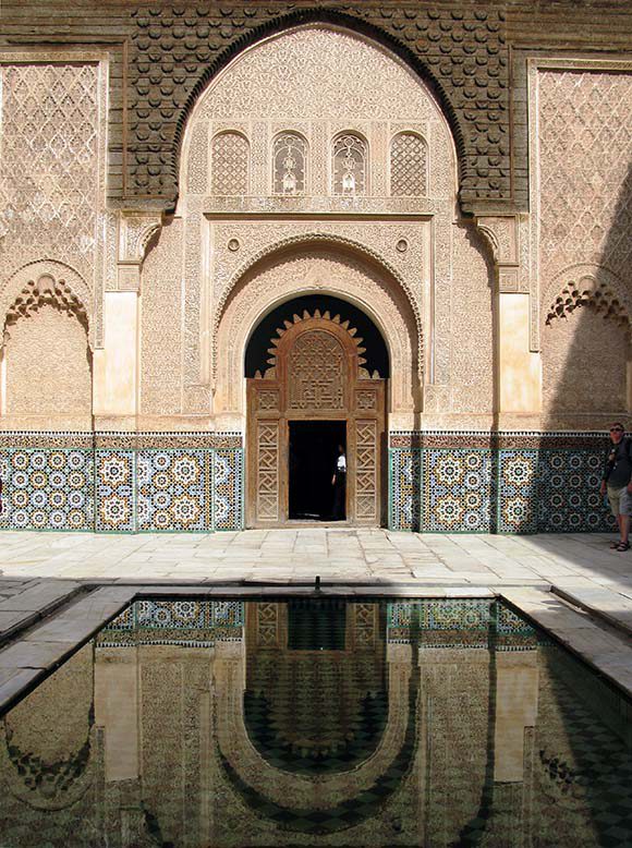 View of a Moroccan building showing a large reflecting pool in front, colorful tiling on the walls, and intricately carved wood around and on the door.