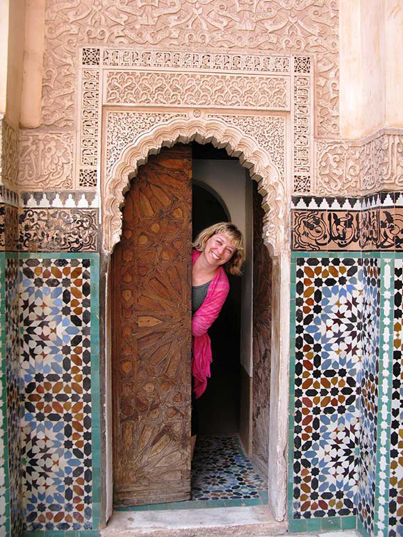 A small doorway in Ben Youssef Madrasa, Marrakech. The wooden doors are carved with a girih pattern of strapwork. The arch is surrounded with arabesques; to either side is a band of Islamic calligraphy, above colourful geometric zellige tilework. Kimball poking her head out from behind the wooden door, wearing hot pink.