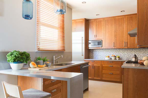 View into a modern kitchen with warm colors of browns, oranges, and tans, reddish-brown wood cabinets, grey countertops, built-in appliances.