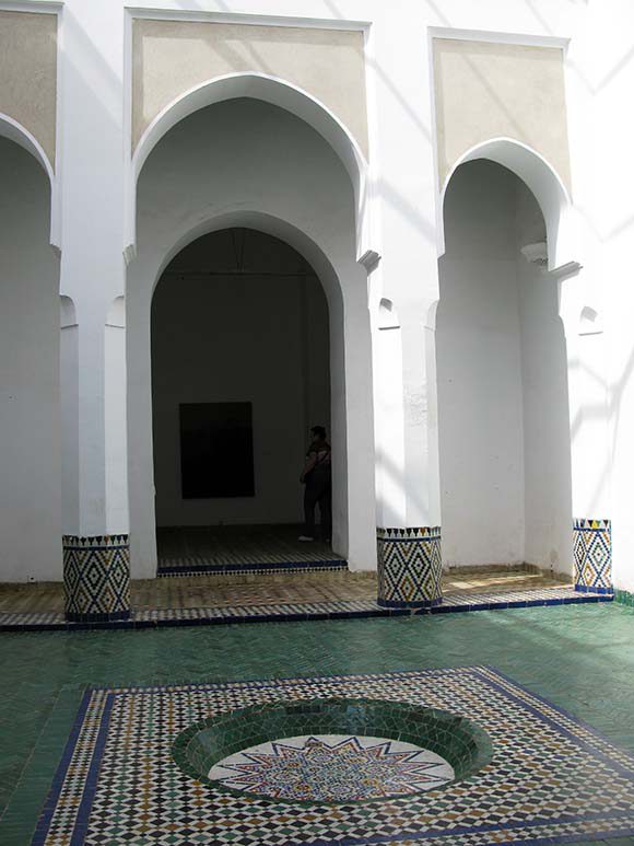 View inside a Moroccan courtyard, with white columns between plain arches, and a sunken tiled water feature in multicolored tiles with sea-green tiles around the outside.