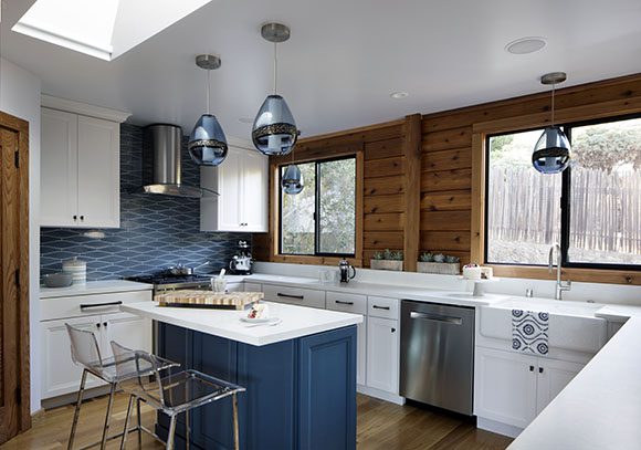 Wood farmhouse kitchen in white, cedar and farmhouse blue cabinetry and backsplash with white quartz countertops, showing a new skylight, dual windows, and multiple blue glass pendant lights.