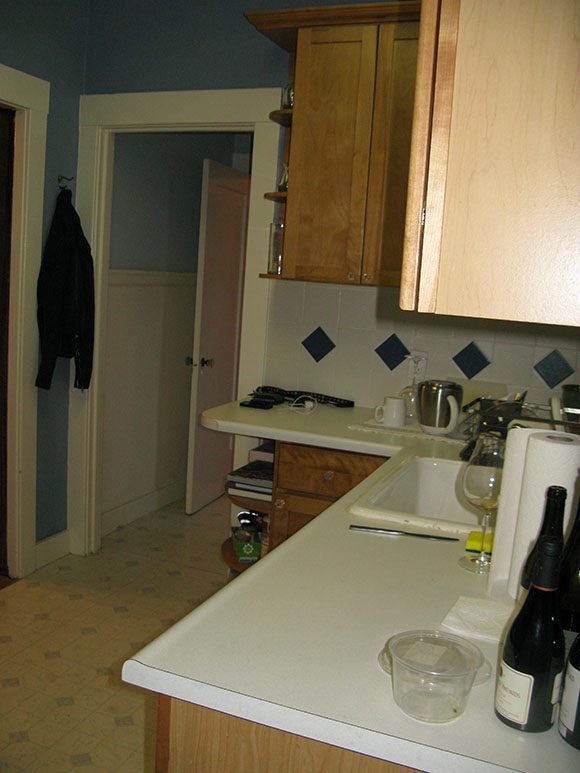 View of L-shaped kitchen countertops, white wall tiling with pine green diamond shape accents as a backsplash, with dated orange wall-hung cabinets, and a door to the left of the space