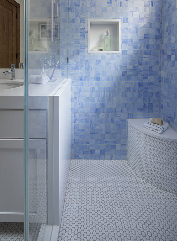 View looking into the glass shower, with bright blue multicolored mosaic tiling on walls, round white penny tile on floor of shower and into rest of bathroom, a curved corner seat to the right, to the left is white quartz countertop, white painted cabinetry and a mirror and lights.