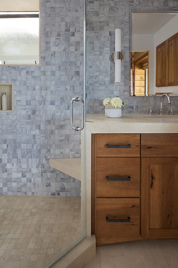 Head-on view into master bathroom, showing glass shower to left at an angle, with grey-blue mosaic tiling on walls, tan tiling on floor of shower, a bit of the angled right corner shower seat in the same marble as countertop and marble flooring, orangey brown wood cabinetry with black metal hardware, lights either side of the mirror.