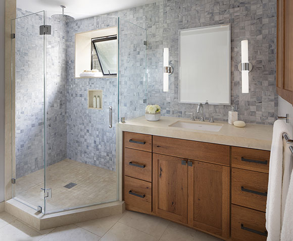 View of a Belmont, CA bathroom, showing glass walk-in shower at left with an open window on the back wall of multi-shade-blue mosaic tiling, to the right of shower, wood cabinetry with black metal hardware, tan countertops and flooring, and sidelights beside mirror.