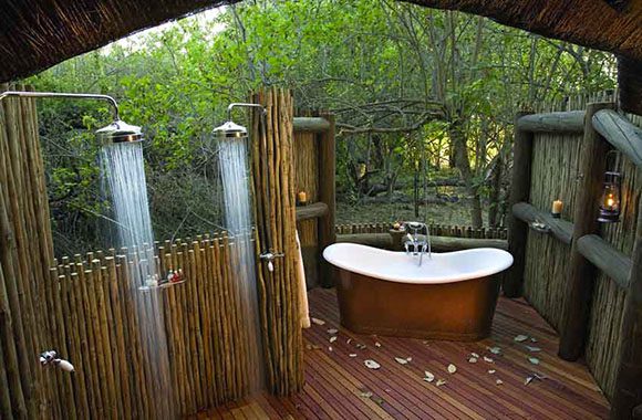 This outdoor bathing area is peeking out from under an arch. We see 2 rain-head outdoor showers flowing, behind bamboo privacy screens. To the right, a copper tub, white enamel inside with centered hand-held nozzle and taps, surrounded by trees and wooden privacy fencing. Reddish wood planking is underfoot in this outdoor bathroom.