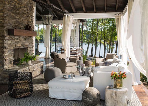 Outdoor living room featuring a rock fireplace surround and metal wood storage caddy, surrounded by billowing white drapes. On a patterned rug sits a sawed wood log side table and a glass side table either side of a white loveseat, hanging lantern lighting overhead, and a gorgeous view out onto a lake.
