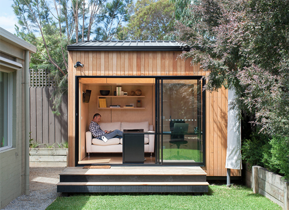 Backyard shed or tiny house used as modern home office and study, two steps up from grass, through sliding glass doors surrounded by blonde wood cladding into a white loveseat, glass desk and dark office chair with computer, 2 bookshelves.