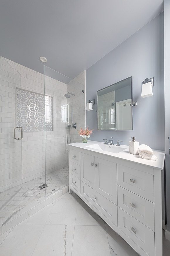 View of bathroom showing pale blue tinted wall and ceiling color, 90-degree angled white porcelain floor tiles, white wood vanity with a white counter surface, silver hardware, and a silver-framed mirror above with silver and white glass sconces either side. To the left is a walk-in glass shower with white subway tiles, a real Calacatta and Celeste marble patterned shower wall niche, complementary angled white porcelain marble-look floor tiles, and silver shower fixtures.