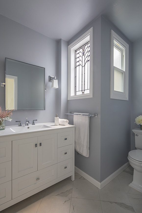 Corner view of bathroom showing white counter surface and white wood cabinetry with blocky silver hardware that matches the silver taps and mirror frame, and one visible of a pair of silver sconces with white glass shades. Next to the light fixture and above the silver double towel rail, are two windows at a 90-degree angle creating a light-filled corner coming in to the space - the window facing the sink is a custom stained glass feature window - and to the right side of the second window sits the toilet in its new location.