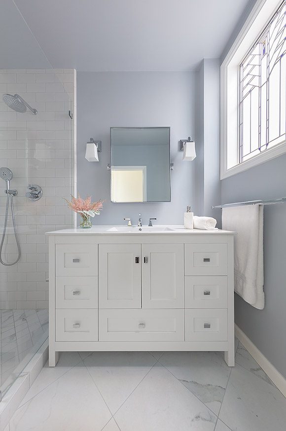 View of upscale bathroom remodel showing pale blue tinted wall and ceiling color, 90-degree angled white porcelain floor tiles, white wood vanity with a white counter and a mirror above with white glass sconces either side. To the left is a shower with white subway tiles and silver fixtures, and to the right is a stained glass window.