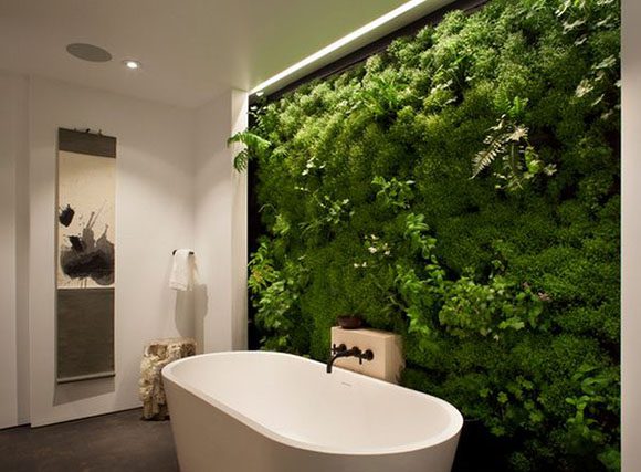 A modern white freestanding tub with black fixtures on a post mounted into a lush green living wall of ferns and mosses is the feature, lit by downlighting. A tall art canvas adorns the side wall, with a towel rail hanging next to the tub.