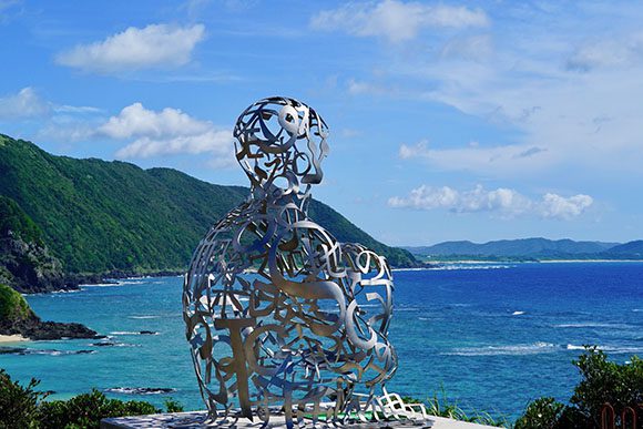 Metal sculpture of a seated human facing the ocean under a blue sky with puffy clouds. The sculpture is made up of words and shapes, by artist Jaume Plensa. To the left are green cliffs, and the water fades from deep blue down to lighter blue as it nears land.
