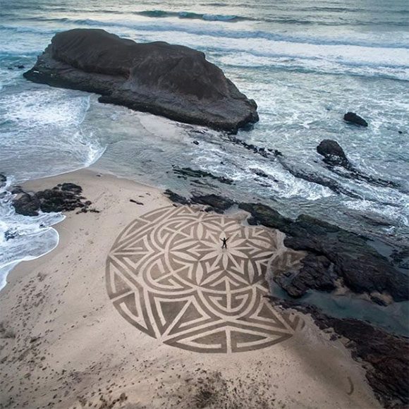 Enormously-scaled geometric circular pattern created on beach sand using only brushes and water, with the artist Andres Amador standing at the center, arms upraised. The pattern is broken by rocks at the edge of the water, between the sand and ocean.