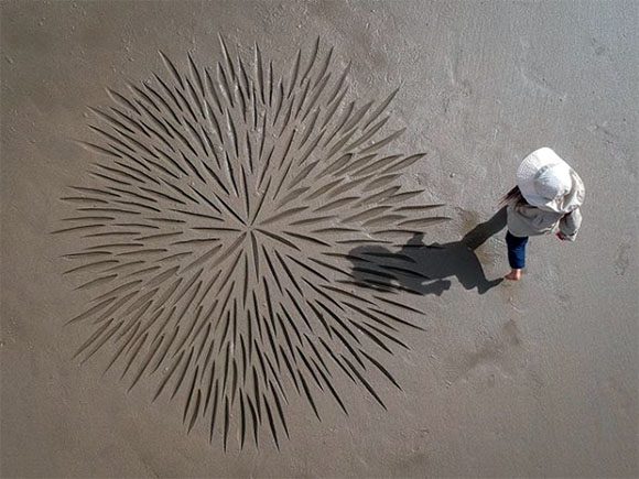 View from directly above, a person walking past a circular central pattern, created with individual long strokes, appearing as if a huge dandelion head landed in the sand for a moment. By artist Andres Amador, who works entirely with brushes and beach sand to create geometric patterns and shapes.