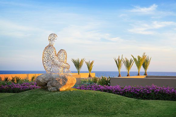 Wire frame sculpture of a human seated on a rock on green grass, contemplating across purple flowers and waving treetops, out towards the sea. Created by artist Jaume Plensa.