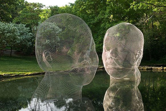 Artist Jaume Plensa creates two silver wireframe heads that appear to float on calm waters, as if listening, by green grasses. The reflections are equally as detailed as the wire sculptures.