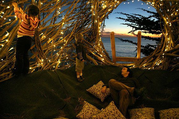 Looking out over ocean waves at orangey sunset, a wooden ladder leads up to a human-scale nest, created by folded tree branches. A child hangs white fairy lights while a man sits among brocade pillows, inside the nest. Artist: Jayson Fann.