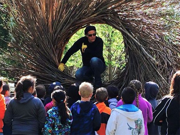 Artist Jayson Fann talks with schoolchildren, seen from behind, about his Nest Creations outdoor art, while seated in the center of the bent branches of one of his nests.
