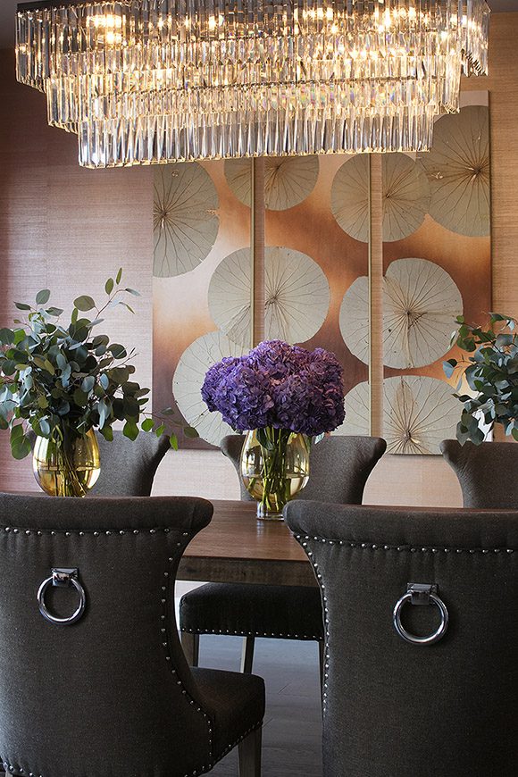 Close-up view of a glamorous transitional dining room showing a thick wooden dining table, dark brown dining chairs with nailheads and a ring detail on the back. Coppery grasscloth wallpaper complements a triptych artwork on the rear wall showing real lotus leaves finished in copper. A rectilinear glass chandelier provides sparkle overhead. Live purple flowers are the centerpiece on the table, with greenery in vases either side.