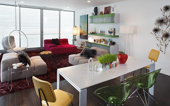 A retro-futuristic living room for a bachelor, with one red and two pink B&B Italia multi-directional sofas on a deep red wine-colored shag rug. To the front is a modern white dining table, two yellow kitchen chairs, and two clear green perspex modern chairs. On the wall behind one of the pink sofas is custom-made shelving and storage, with bright green wood and clear perspex sliding panels. An Eero Aarnio bubble swing chair hangs just in front of a large set of windows, with the projector screen hung just above, ready to pull down for a movie projected from the ceiling.