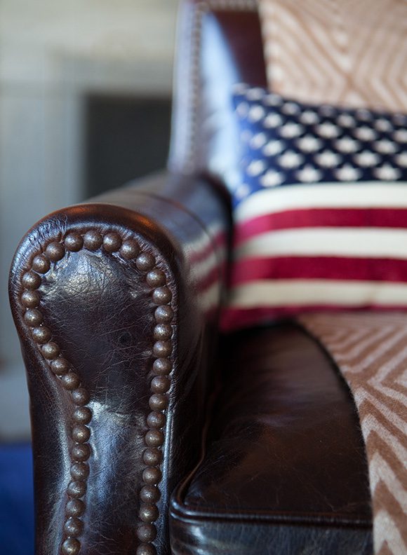 Close-up image of the right arm of an aged dark leather club chair with round nailhead edging. On the seat is a folded patterned blanket, and on top of that, slightly out of focus, an American flag pillow rests against the back of the chair.
