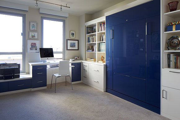 A hardworking home office converts to a comfortable guest getaway with this convertible bed, hidden behind a large custom-made blue wall cabinet. A white desk chair and a blue-and-white desk sit in front of a window with roller blinds. The light colored carpet helps with sound attenuation.