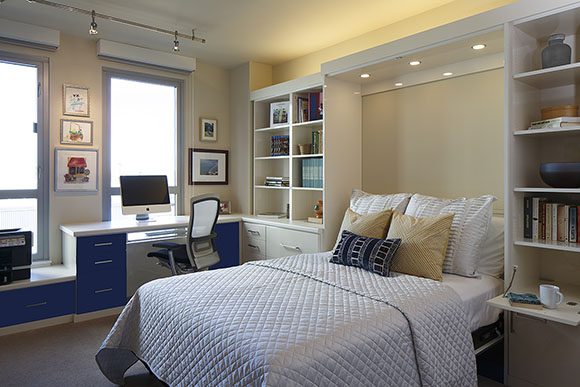 A hardworking home office converts to a comfortable guest getaway with this custom-made Murphy bed. Uplighting spreads light while downlighting provides a handy reading light. A shelf folds down to create a bedside table. The bed itself is dressed in soft white, yellow, and blue pillows, with an icy blue comforter. A white and silver desk chair is neatly tucked under the blue-and-white desk in front of the window with roller blinds up.