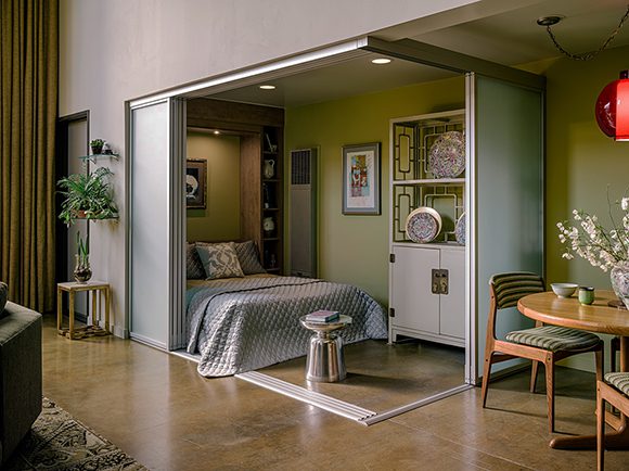 View into a corner room with sliding perspex doors folded back to reveal the convertible bed dressed with fresh green and white linens. The two slipper chairs have been removed, and the side table is now perfect for books and magazines. A vintage dining set in blonde wood and green striped fabric shows to the right. The flooring is light-colored wood, and the walls are a light green shade.