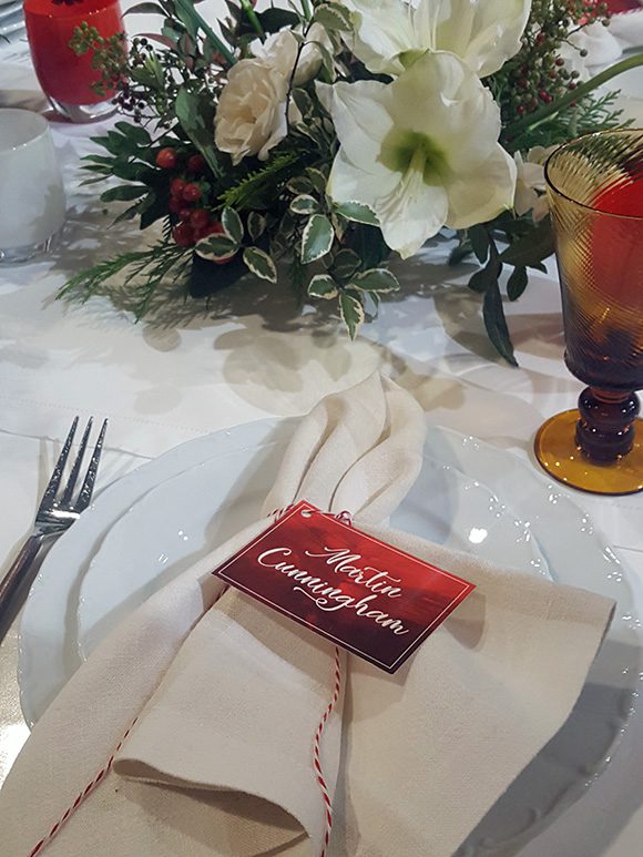 Closeup view of a holiday dining place-setting. At the front of the image, two stacked white china plates are placed with a white linen napkin, tied with red and white twisted string to hold the napkin. White-lettered red namecard is attached to the string. A fork is visible to the left, and a tan fluted glass is just showing in front of the white, red and green floral centerpiece arrangement, which is cut off at the top of the image.
