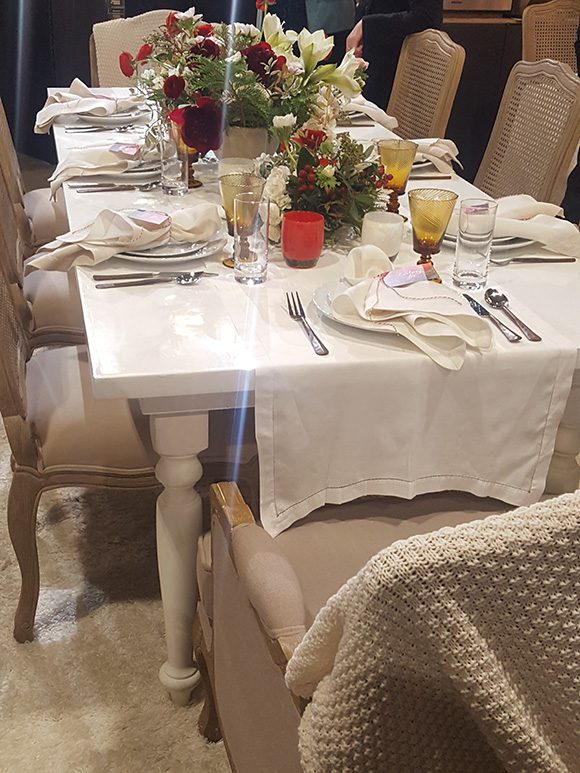 View of a rectangular dinner table from the short end, with a white textured blanket over the back of the head chair showing bottom right of image. Table is light-colored with wooden legs and white marble top. Chairs are very light taupe with a pierced pattern cane back and velvet fabric seat. The table runner is white with white linen napkins topping the formal place settings for 6 people. Tan colored fluted glasses are next to clear water glasses. The center of the table is filled with red, white and green plants and flowers in a festive arrangement.