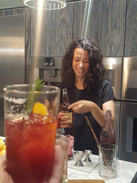 Holding an example reddish mixed drink in hand, filled with ice, in the bottom left of the image. The rest shows a woman making a shrub cocktail drink, holding a bottle of vinegar, with several glasses, bottles, and metal measurers on the counter in front of her. The kitchen space behind her is a modern design with textured wood grain and stainless steel appliances.
