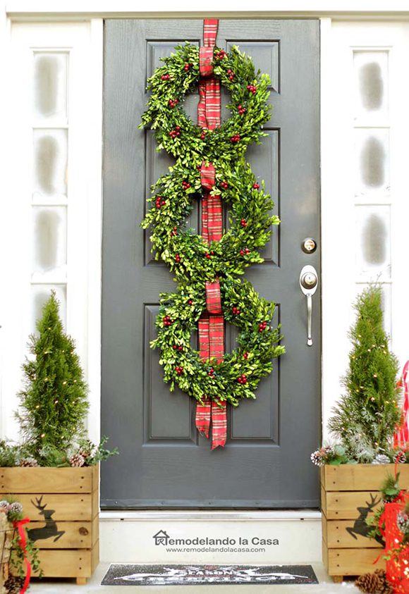 Image of a dark grey to black front door decorated with 3 vertical wreaths in bright green with red berries, hung on red and gold ribbon. Either side of the door are small planter boxes containing tiny boxwood trees. The boxes are decorated with leaping reindeer or stags burned into the wooden planks, for the holidays. The full-length windows either side of the door are frosted to resemble snow.