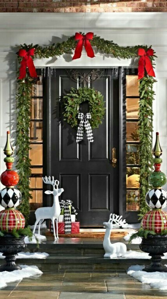 Image of a residential front entrance with a black front door and low, dark stone steps over tan and grey flagstones at the sidewalk level, icy snowdrifts at the edges, and white deer statues on two steps. Either side are statues of 3 balls or globes balanced, the bottom ones in plaid, the middle ones in black-and-white harlequin diamonds, and the highest in red or green, topped with gold spires. Behind the standing deer is a small stack of 3 gifts or packages wrapped with harlequin diamond ribbon that matches the middle globes. Swags of evergreen are tied with wide red ribbons over the door on a white door surround and hanging above the sidelight windows, and a matching evergreen wreath hangs in the center, with a harlequin bow to finish it off nicely.