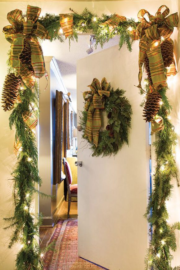 This image is of an apartment door, partially open to reveal a sliver of the inside hallway and a chair beyond. Back at the main hallway exterior, large swags of evergreens, huge pinecones, and small white Christmas lights are tied with two oversized striped green-and-gold bows at either corner. An evergreen wreath with matching bow hangs in the center of the plain white door.