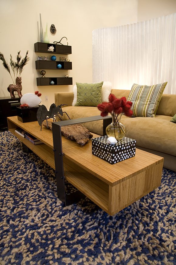 Angled image of a living room, showing muted eggshell paint on walls, blue and white speckled rug, black metal support on a wooden coffee table dressed with flowers in a vase on top of a blue and white polka dotted box along with other curios, a tan sofa is just behind the coffee table. Brown floating shelves on the wall at the rear left are scattered with sculpture and collectibles but plenty of open space left between them, and an opaque white folding screen stands behind the sofa, softly diffusing light into the room. Striped and patterned pillows rest on the sofa.