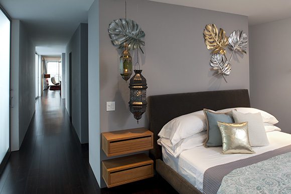 View in a bedroom, with a long hall to the left, the bedroom to the right, painted in a warm gray paint color, with a dark headboard and white linens on the bed, topped by 3 different colors of accent pillows – blue, gold, white. Metallic palm fronds adorn the walls. 2 pendant lamps hang just over the floating drawers that act as bedside table in a warm brown.