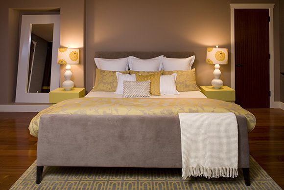 Low lighting from a pair of bedside lamps in white with yellow flowers sit atop a matched pair of bright yellow bedside tables. The gray suede footboard and headboard have a lilac hue to them, offset by multiple layers of white and yellow pillows with gray patterns. A yellow and silver floral comforter repeat the tones from the interlocking patterned rug that protects your feet from the cold of the shiny cherry-colored wood flooring. A door to the right and a mirror to the left complete the balanced look of the room.