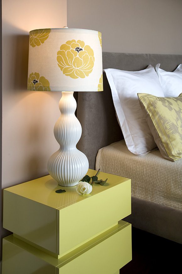 Close-up view of the daffodil-yellow lacquered bedside table, a modern square stacked on top of another square, without drawer-pulls or visible hardware. The white triple-bubble-shaped lamp is topped by a lampshade decorated with graphic yellow flowers with a purple center. Textures on the bedlinens and pillows are visible. A single white rose is laid on the table by the bed.