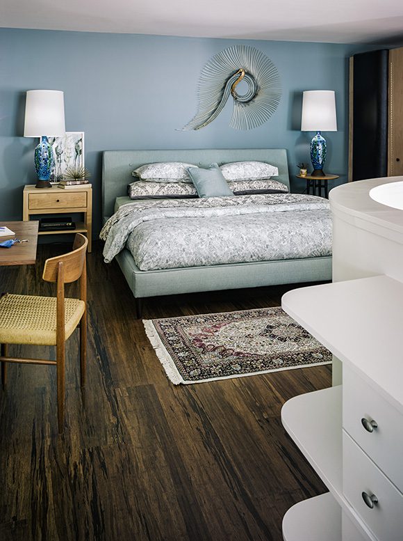 A sky blue wall paint color is the backdrop to this restful bedroom, showing 2 ceramic blue lamps on blond bedside tables, either side of a sea-blue-green headboard and bedstead, with white and gray patterned sheets and 2 stacks of pillows, and a matching blue accent pillow in front. The dark wood floor contrasts against an oriental rug at the foot of the bed.