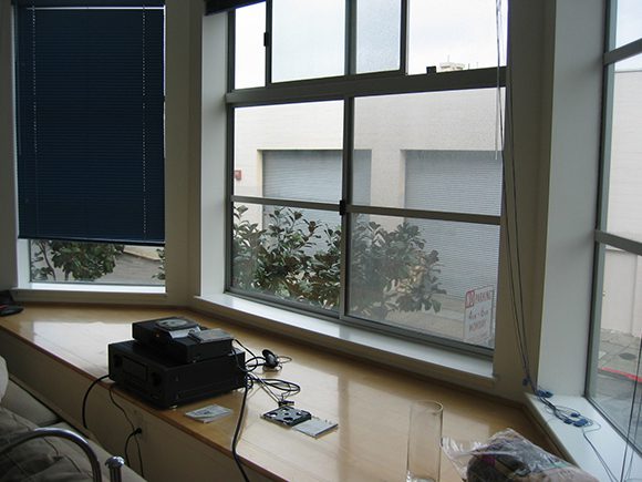 Large bay window with simple wood framing, a single blind pulled down over the left window, and some video game components rest on the large windowsill finished in glossy light-colored wood.
