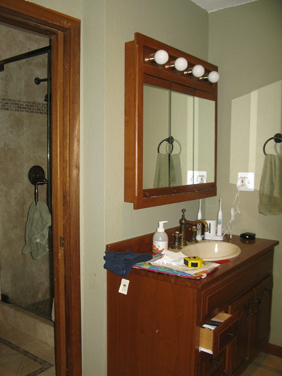 In this homeowner's 'before' photo, the shower was previously separated from the sink by a sliding door. Very dark wood cabinetry and countertops contrasted with the light-colored sink. Above that was an 90s "glamor" line of lights consisting of round bulb light fixtures above a triple-mirrored prescription cabinet. This split room did't make good use of the floor space, and the greenish-grey wall color didn't help put you at ease.