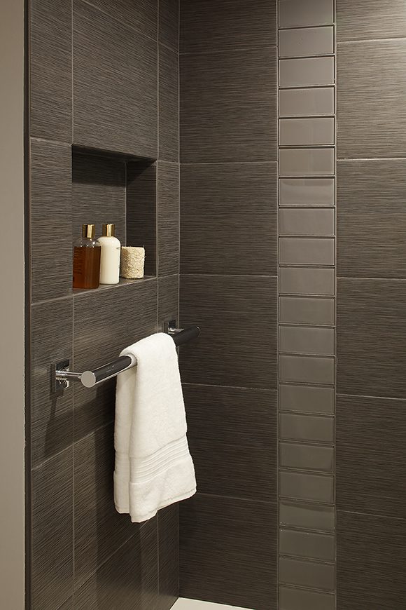 Closeup of a corner of a contemporary bathroom with a white towel folded neatly, hanging on the silver towel rail on the left wall inside the shower. Above the towel rail is a recessed niche is styled with luxury shower product bottles, and a candle. The subway wall tiles are dark brown with a greyish horizontal striated texture. A single vertical row of flat reflective brown tiles runs from top to bottom on the back wall. A tiny strip of the grey wall paint shows at the far right of the image.