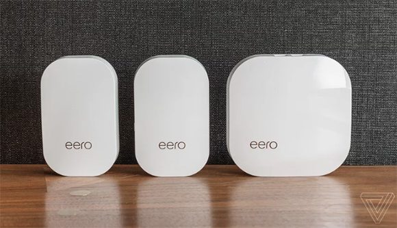 Two Eero pods and a base station are pictured sitting upright on a dark wood shelf, in front of a grey textured wall finish. The Eero units are all white, square with rounded corners. The base station is completely square, while the pods are more rectangular. There are no buttons visible in this view, but the base station shows a slight area where plugs can be inserted. They are made of a sleek shiny plastic finish, and look very modern. They bear the logo for Eero, which is written all in lower case letters in a sans serif font.