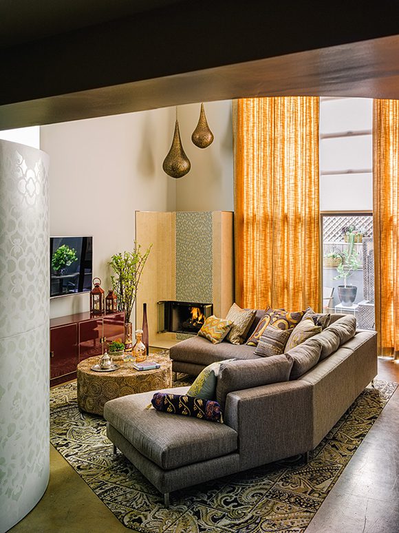 View into a Francisco loft contemporary living room, which mixes a midcentury modern sofa with Moroccan influences in a patterned ottoman used as a coffee table, and teardrop-shaped brass pendant lamps. Full height gold curtains filter sunlight into the space and a yellow and green patterned rug anchors the living area in front of a wall-mounted TV over a midcentury sideboard used as media storage.