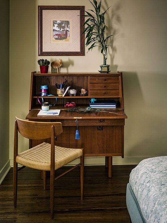 Gold wall paint color sets off a vintage mid-century modern secretary desk in a warm brown wood, with a yellow-seated vintage chair, also in the same warm wood tones. A piece of bamboo sits atop the desk, next to art hung on the wall. A hint of blue and white bedding at the bottom right corner indicates a bed.