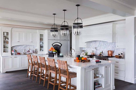 East Coast meets West Coast in this Cape Cod-inspired kitchen, mostly in white finishes, with black metal accents as seen on the three pendant lights. Wooden counter seating spans the kitchen island, which is a major design element.
