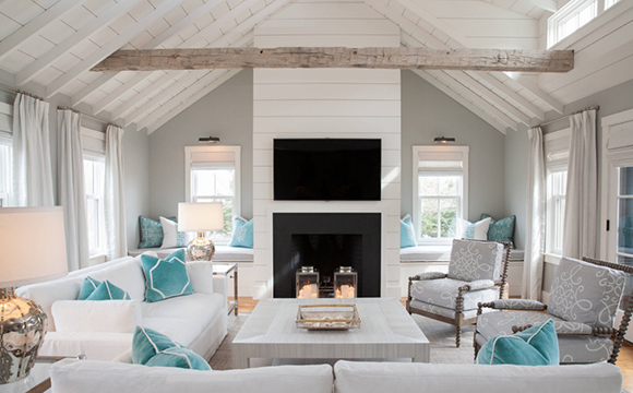 View of a living room with a single exposed beam at the ceiling, off-white wall color, curtains, and ceilings, grey and tan toned furniture, with just a few pops of turquoise color brought in with throw pillows. A black fireplace and TV screen above it are the darkest elements in the room, with everything else being soothing shades of neutral colors.